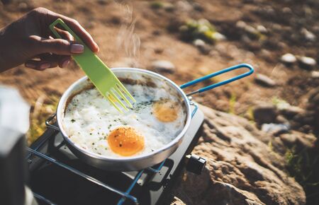 person cooking omelette eggs in nature camping outdoor, cooker prepare breakfast picnic on metal gas stove, tourism recreation outside; campsite lifestyleの写真素材