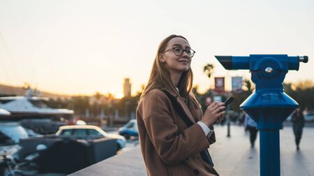 Traveler woman holding mobile phone. Girl tourist in glasses using online smartphone technology. Digital communication on sunsetの写真素材