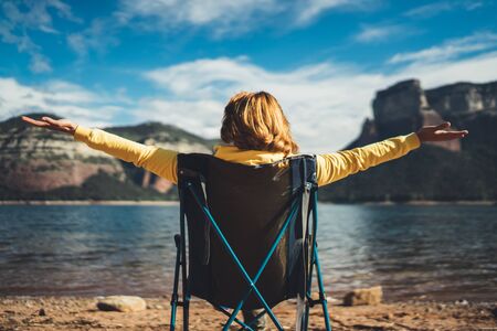 happy tourist traveler sits with raised hands, girl hiker enjoy mountains lake breathing of fresh clean air in trip in spain, relax holidayの写真素材
