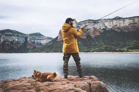 beard fisherman hold in hands fishing rod, man enjoy hobby sport with pet dog on lake, person catch fish on river on background mount,  fishery conceptの写真素材