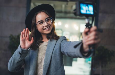 Female vlogger recording with digital camera. Smiling young woman taking selfie video on light night city. Traveler blogger shooting video for shoot social mediaの写真素材
