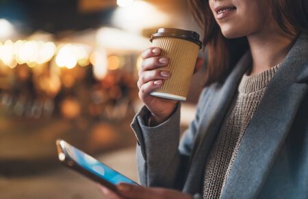 Smile girl drink coffee outdoors using mobile smartphone on background bokeh light in night city, womn hold in hands ecology disposable cup of tea , online  internetの写真素材