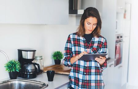 workplace at home, girl study cooking process through Internet  technology on tablet and drink coffee in kitchen, girl person holding computer on background interior cuisine, social distance conceptの写真素材