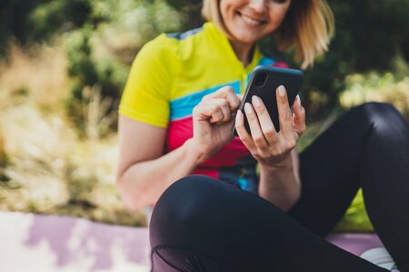 Young smile woman communication mobile phone on nature. Girl using smartphone online internet technology after training fitness in summer park. Tourist relax after exercising sport outdoorsの写真素材