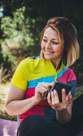 Fun girl using smartphone online technology after training fitness in summer park. Tourist communication on cellphone relax after exercising sport outdoors, healthy lifestyle concept. Young smile woman calling on internetの写真素材