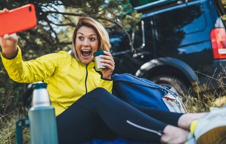 Girl joy showing teeth call on mobile phone drink tea in summer forest. Happy tourist communication take selfie on cellphone while traveling auto. Isolation woman using smartphone have fun emotion outdoorsの写真素材