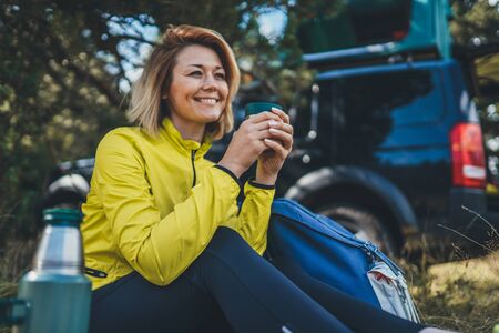 Girl smiles enjoys breakfast outdoors summer nature, tourist hold hand mug  warm tea during recreation trip, hiker laughing drink coffee from  in green forest. Happy tourist relax while traveling autoの写真素材