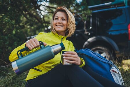 Girl smiles and enjoys summer nature hold hand mug of warm tea during recreation trip, hiker laughing showing teeth drink coffee  in green forest. Happy tourist relax outdoor while traveling auto tourismの写真素材