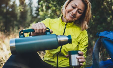 Girl smiles drink coffee  on nature after training, woman hold hand mug of warm tea outdoors, hiker enjoys breakfast rest in green forest. Tourist relax while travelingの写真素材