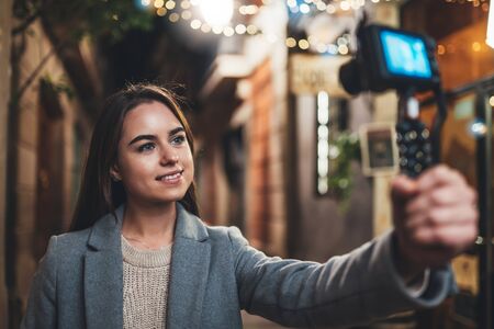 Female blogger shooting video for vlog social media with digital camera. Smiling woman vlogger taking photo selfie on background light night city illuminationsの写真素材