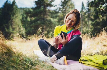 Girl laughing using smartphone technology after training fitness in park. Tourist relax after exercising sport outdoors, healthy lifestyle concept. Young smile woman communication mobile phone on nature, online internetの写真素材