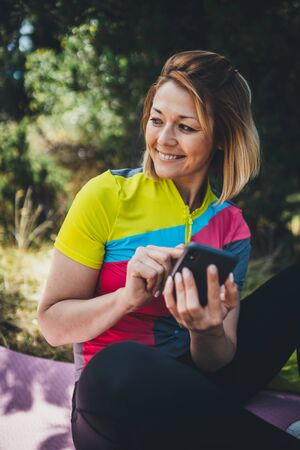 Happy girl using smartphone online technology after training fitness in park. Tourist laughing communication cellphone on nature, healthy lifestyle concept. Young smile woman calling on internet outdoorsの写真素材