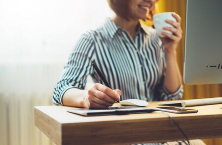 female hands graw on portable tablet at home interior, smile woman drink coffee working at office workplace with digital stylus on computer,  hipster manager using device pen, social isolation work processの写真素材