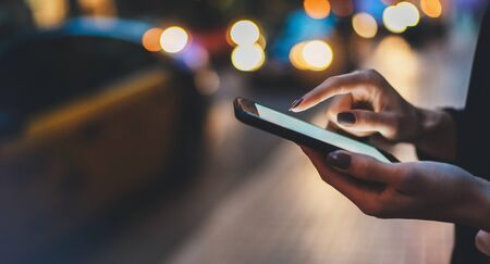 Young woman with smartphone using application for calling taxi online walking on city street night, hipster girl  with internet connection texting friend via mobile phone on background bokeh color lightの写真素材