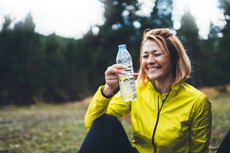 Fun girl quenches thirst after fitness. Smile person laughing drinking water from plastic bottles relax after exercising sport outdoors, woman isolation training in forest on nature, healthy lifestyle concept. Young sportwoman drink water after joggingの写真素材