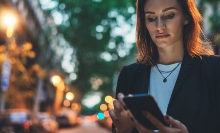 Confident young woman in black suit working documents using smartphone on background lights evening city, female person calling on mobile phone outdoors in the nightの写真素材