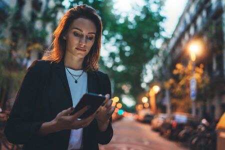 Young stylish business woman writes message on smartphone walking evening city street, hipster tourist traveling in european town and planning trip using mobile phone