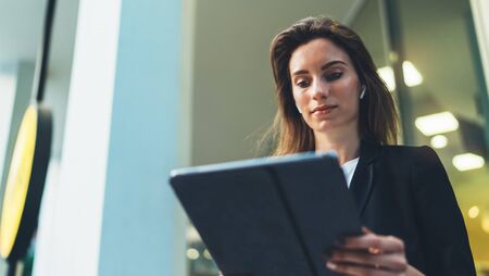 Successful businesswoman using tablet and wireless earphones outdoors near his office background lights, portrait young woman professional manager working on pc computer near window skyscraperの写真素材