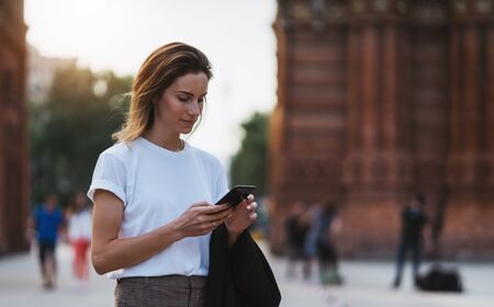 Portrait of beautiful traveler woman reading message on smart phone walking arc de Triomphe of Barcelona, tourist girl using mobile phone in sun summer day park outdoorsの写真素材