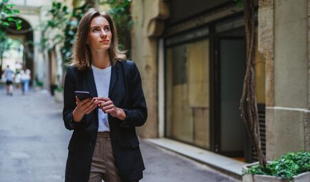 girl looks up at the sights of old European city and reads about them online in a mobile phone,  female manager in a business suit enjoys a vacation while walking along the green streets of Barcelonaの写真素材
