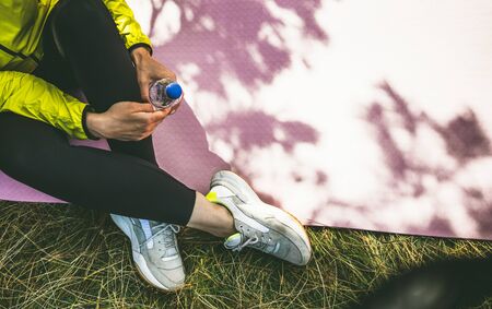 Top view girl  with bottle of water relax after exercising outdoors on yoga mat, isolation fitness woman in sneaker stretch training outside on green grass. Empty space for your textの写真素材