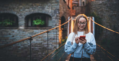 tourist girl with glasses and camera stands on suspension bridge of old historical city uses smartphone to plan evening walk, cute blonde hipster looks at  mobile phone screen enjoying weekend vacation in summerの写真素材