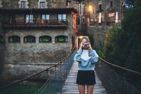 tourist girl with glasses stands on suspension bridge on background historical city and takes photo...