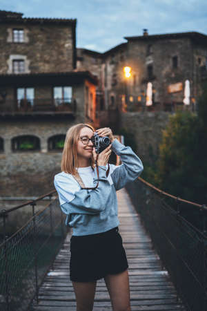 blonde tourist girl stands on suspension bridge walking old historical city and takes photo with retro camera, cute young hipster enjoying a weekend vacation in summer uses photocamera to take picture of her journeyの写真素材