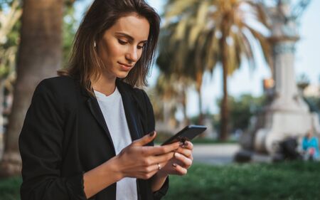 Woman walking and chatting with friends outdoor in city park, Close up of attractive business woman typing on touch screen of cellphone while standing outdoorsの写真素材