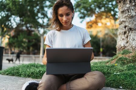 caucasian hipster girl using tablet outdoors, cheerful young freelancer working on touch pad at green park,  female student studying sitting grass with laptop computerの写真素材