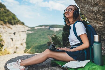 young girl tourist with backpack enjoying summer holidays listen music in headphones and looks online map on laptop outdoor, fun woman copywriter with glasses working on digital tablet device on backdrop landscapeの写真素材