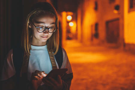 front view portrait of young girl tourist with reflection of glare in glasses from mobile phone screen uses Internet technology at night on street of old historical city background of evening lights, copy space for your textの写真素材