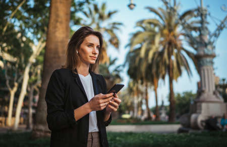 elegant girl in suit holds a smartphone in hand and looks into distance while walking in a Park in Barcelona, girl Ð¼anager checks chat with colleagues on mobile phone after office workの写真素材