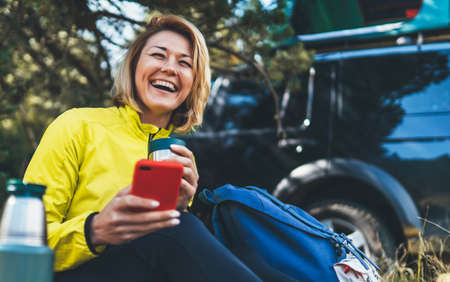 Happy auto tourist laughing talking on cellphone drinks tea while traveling. Young woman smile showing teeth calling on internet mobile phone summer green forest. Joy girl using smartphone have fun emotion in outdoorsの写真素材