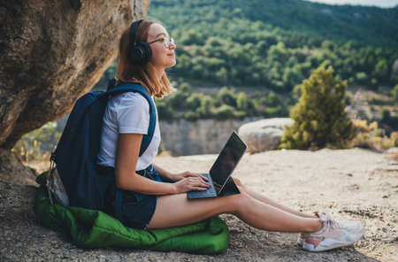 blonde girl after walk in mountains rests listening to music in headphones typing on a laptop in nature, traveler woman plans route in digital tablet looks at online map for walkの写真素材