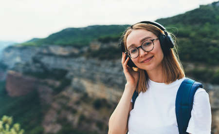 positive young traveler with long blonde hair and hipster glasses listens to favorite music in wireless headphones smiles while hike walking trip through the natural mountain landscape on summer vacationの写真素材