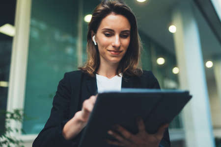 Successful female banker using tablet and wireless earphones outdoors near his office background lights, portrait young woman professional manager working on touch pad near skyscraper in evening cityの写真素材