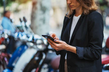 modern woman in business suit holding in hands smartphone uses app scooter rental service outside in city,  female manager looks at map on her mobile phone to rent scooter for quick trip to office, Ñoncept of urban transportの写真素材