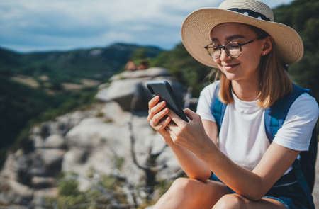 tourist woman with hipster glasses and summer hat using mobile phone device in hands internet technology to enjoy hiking walk on top of mountains during freedom vacationの写真素材