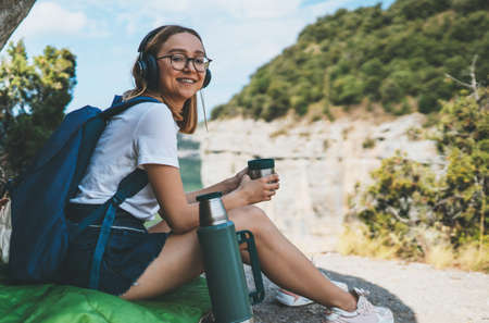 young girl smile traveling in mountains drinking coffee from flask enjoying summer trip, fun female hipster tourist with glasses relaxing in nature vacation listening music with headphones, copy space for textの写真素材