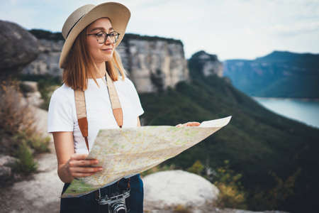 tourist girl in summer hat holding in hands map resting during trip in mountains landscape, hipster woman in glasses plans route resting in nature in open air, empty space for textの写真素材