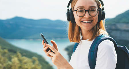 portrait smiling traveler blonde girl with hipser glasses and backpack using mobile phone device and listens music for headphones via Internet technology to relax hiking walk on top mountains outdoors, copy spaceの写真素材