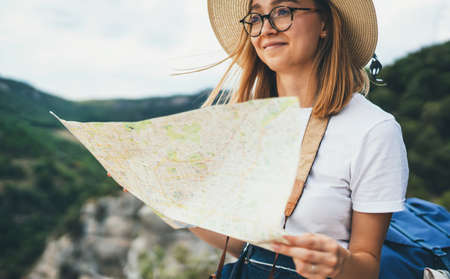tourist girl in hat holding in hands map and plans route leisure in nature during summer vacation on background green mountains landscapeの写真素材