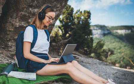 positive young millennial girl with headphones sitting outdoors in mountains working from distance using laptop, beautiful tourist girl with glasses having an online video meeting via computer outdoorの写真素材