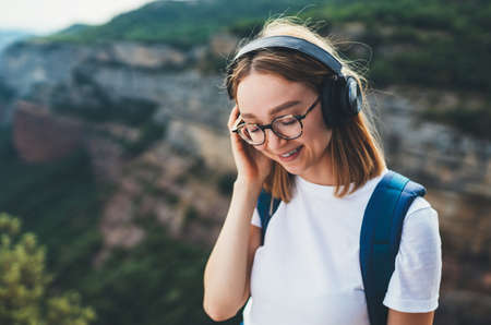 relaxed young traveler woman hipster meditates with eyes closed listening to favorite music on wireless headphones while standing on top of mountain and enjoying freedom and clean airの写真素材