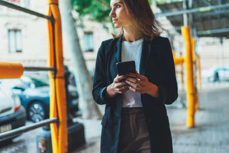 successful businesswoman listens to financial news on her wireless headphones and checks email on smartphone during lunch break outside officeの写真素材