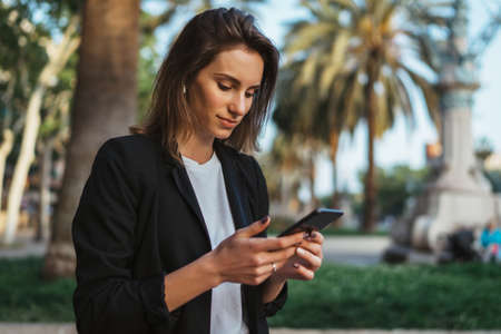 Woman walking outside in city park and hold in hand mobile phone chatting with friends, businesswoman typing on touch screen of cellphone while internet technology outdoorsの写真素材