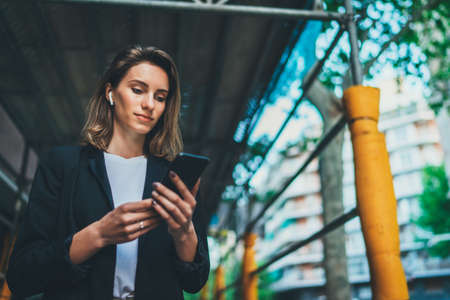 businesswoman listens to financial news on wireless headphones and checks email on smartphone during lunch break outside office, manager using cellphone working in city outdoorsの写真素材