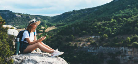 girl traveler in hat resting during vacation in nature and plans route online map in smartphone, tourist girl uses mobile phone while enjoying vacation on background of panorama mountain landscape copy spaceの写真素材
