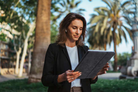 successful young business woman using digital tablet while walking in summer Park barcelona, hipster girl in black jacket holding touchpad looks at computer screen on a city streetの写真素材